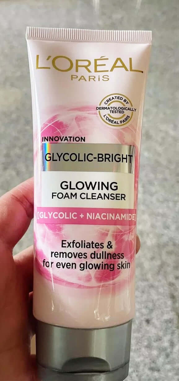 A hand holding a pink tube of LOreal Glycolic Bright Cleanser showing the front of the product packaging.