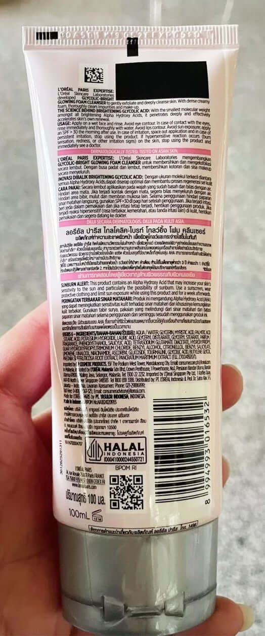 A hand holding the back of a LOreal Glycolic Bright Cleanser tube showing the ingredient list, barcode, and usage instructions.