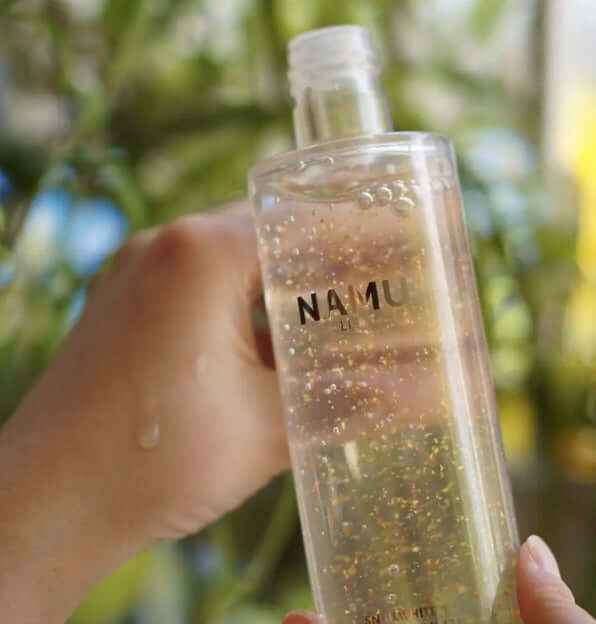 Hand holding a bottle of Snailwhite Gold Essence with visible 24K gold flakes, showcasing skin care benefits.