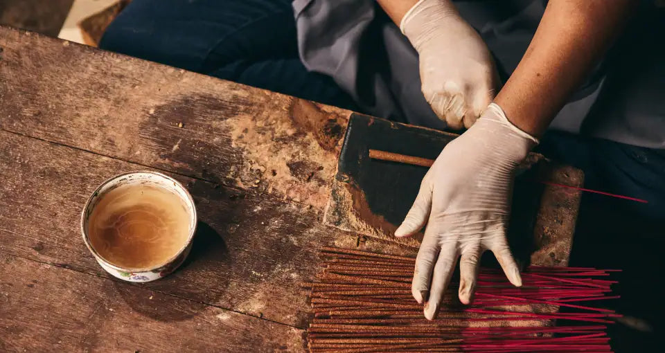 Artisan preparing Noppamas Incense Sticks on a wooden table with a cup of coffee nearby.