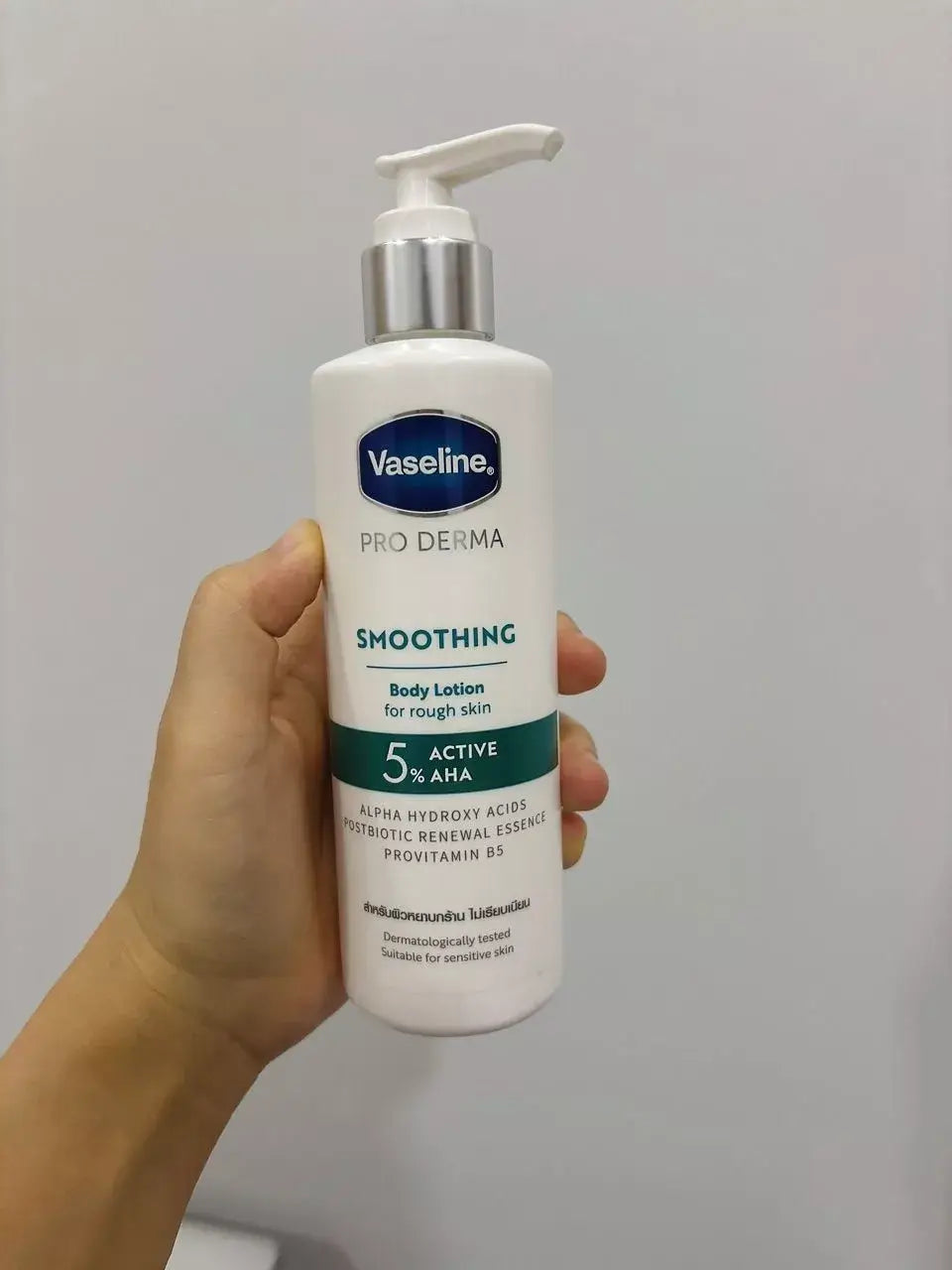 Hand holding Vaseline Pro Derma 5 AHA body lotion bottle in front of a white wall.