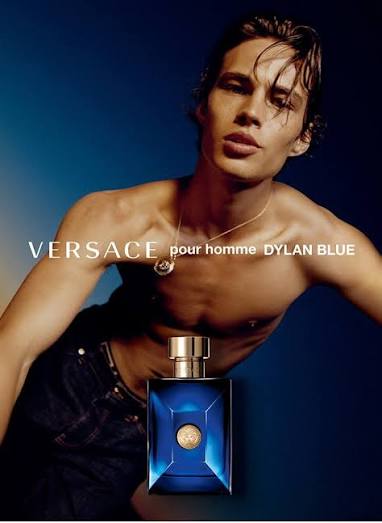 A shirtless man in a black tank top and jeans holding a bottle of Versace Dylan Blue Pour Homme EDT.