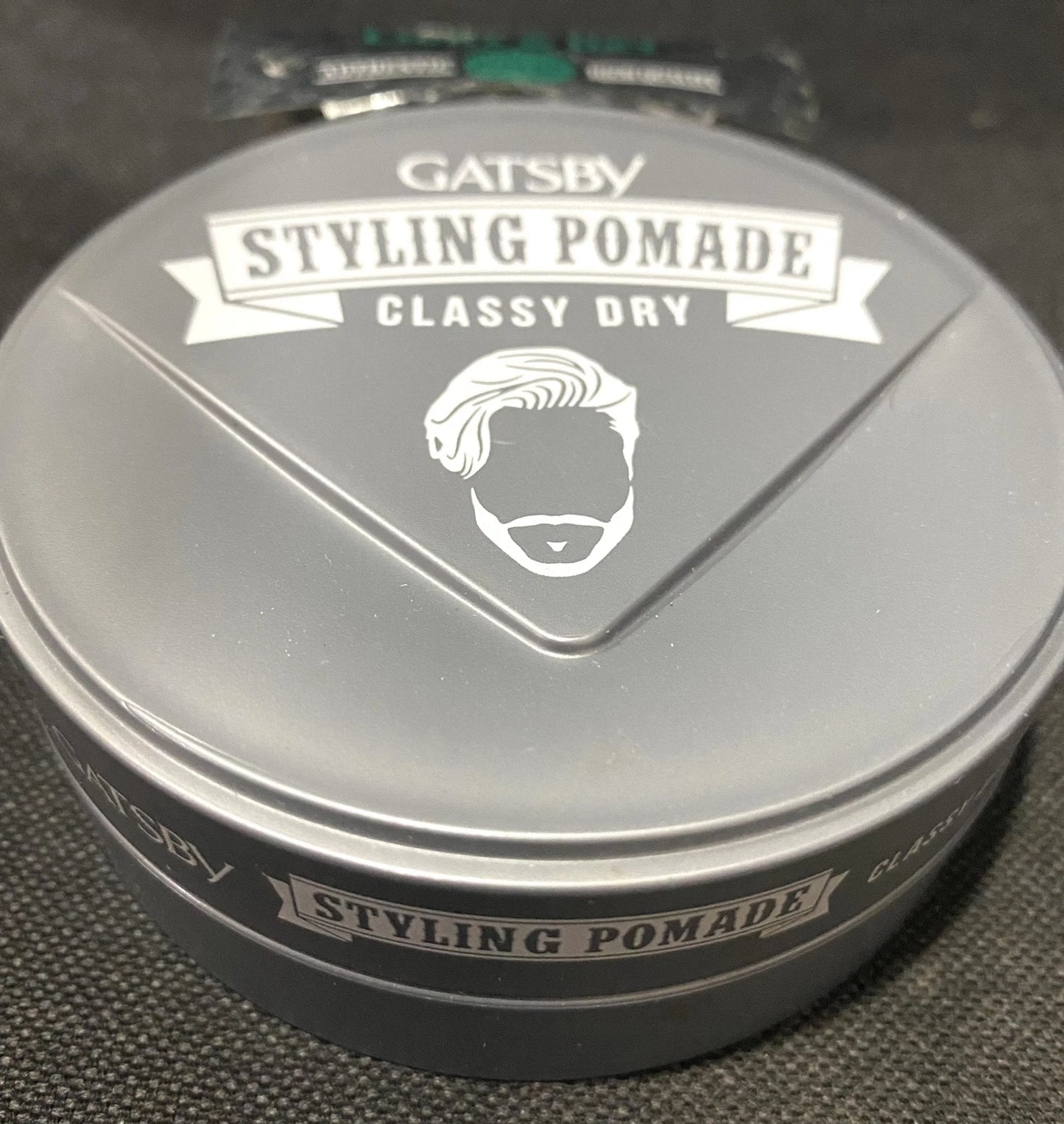 Angled view of the GATSBY Classy Dry Pomade jar and lid with white ribbon text.